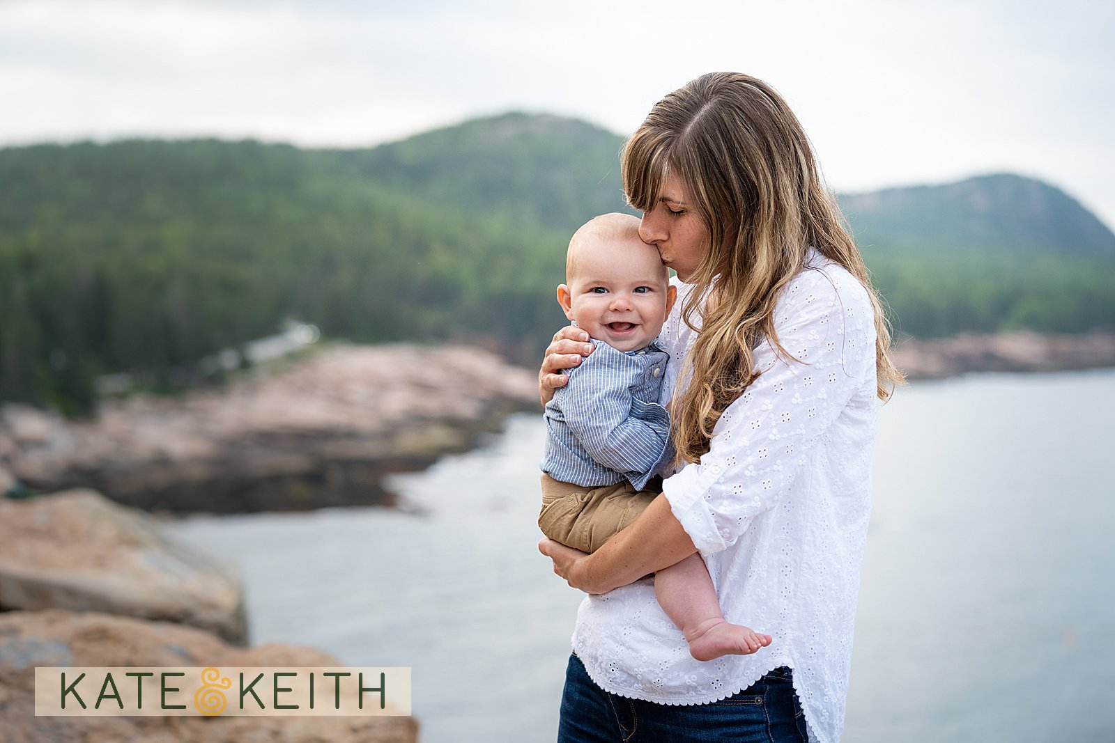 mom kissing her smiling baby with Acadia National Park mountains and ocean in the background