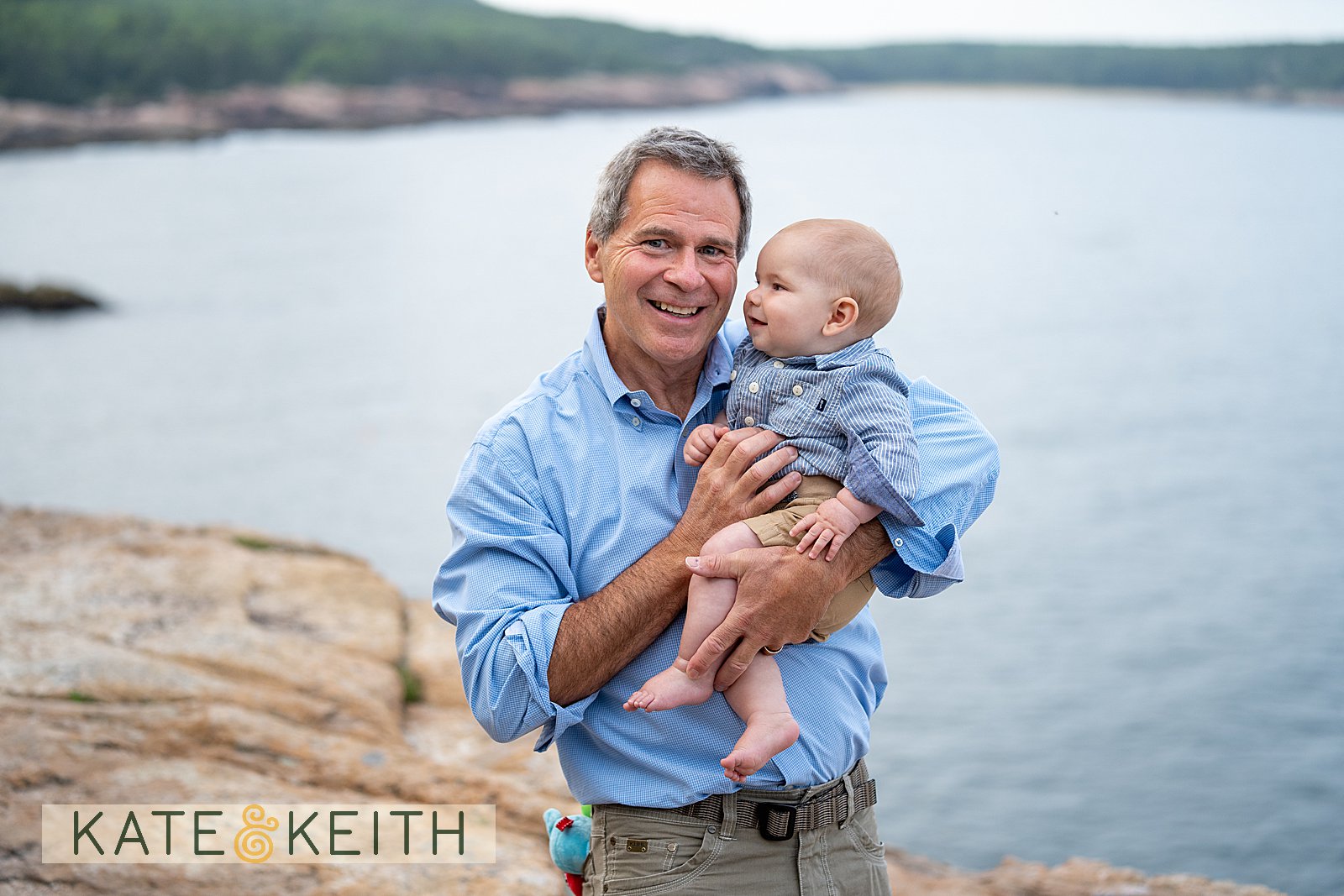 Grandpa smiling and holding his grandson who is looking at him, posing in Acadia National Park