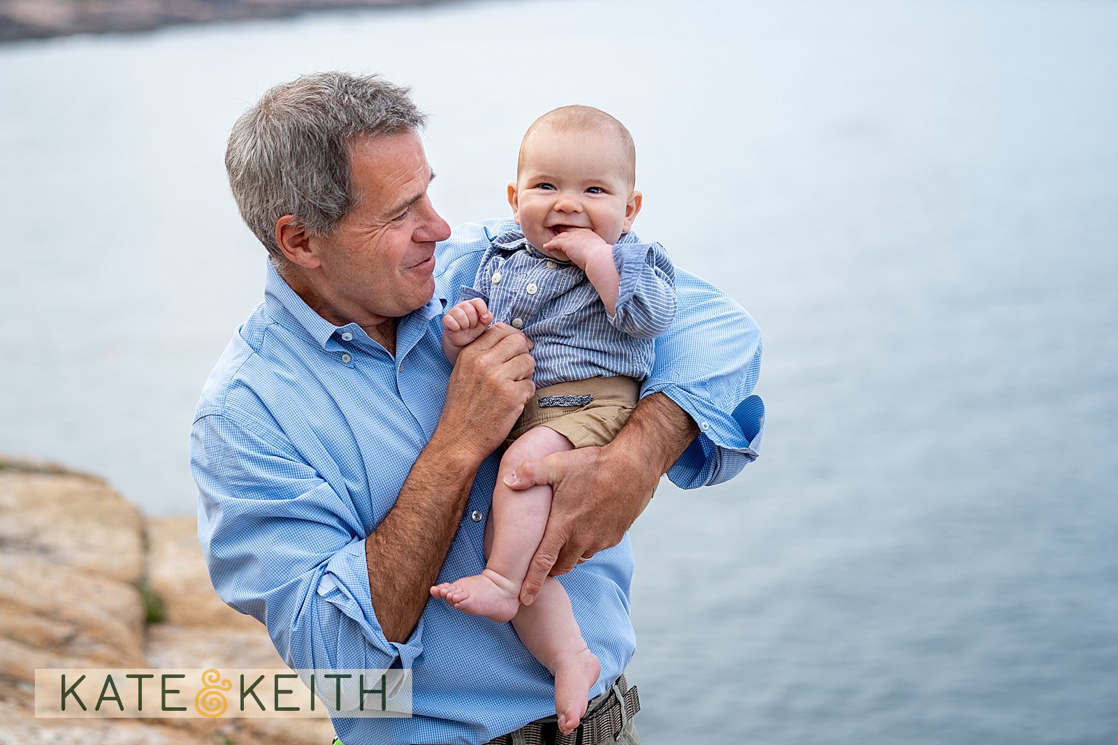 grandpa holding a smiling baby above the Maine coastline