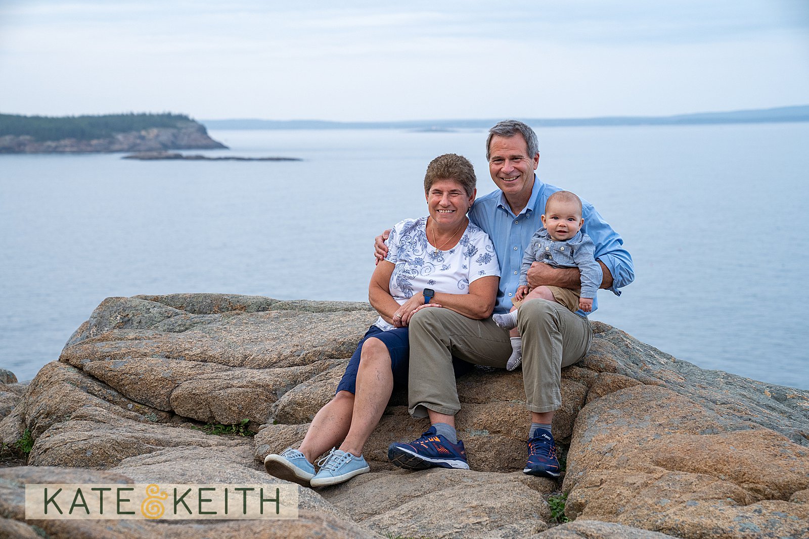 Grandma, Grandpa and baby sitting on the rocks of Acadia with the Atlantic in the background