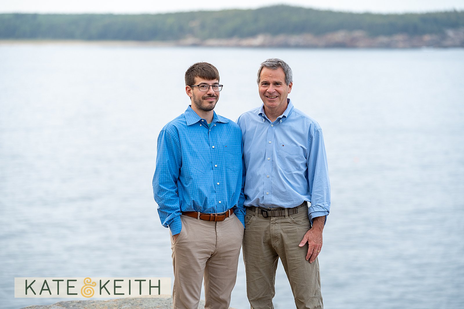 adult son and dad posing on rocks with Acadia National Park as a backdrop
