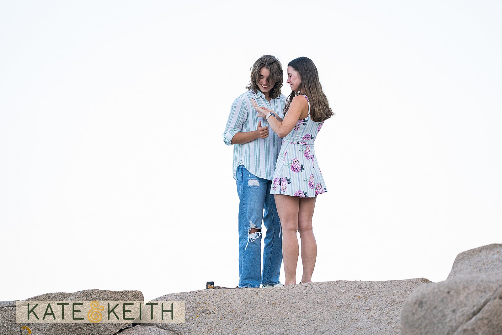 two woman, newly engaged in Acadia National Park, standing on a rock looking at the ring on her finger