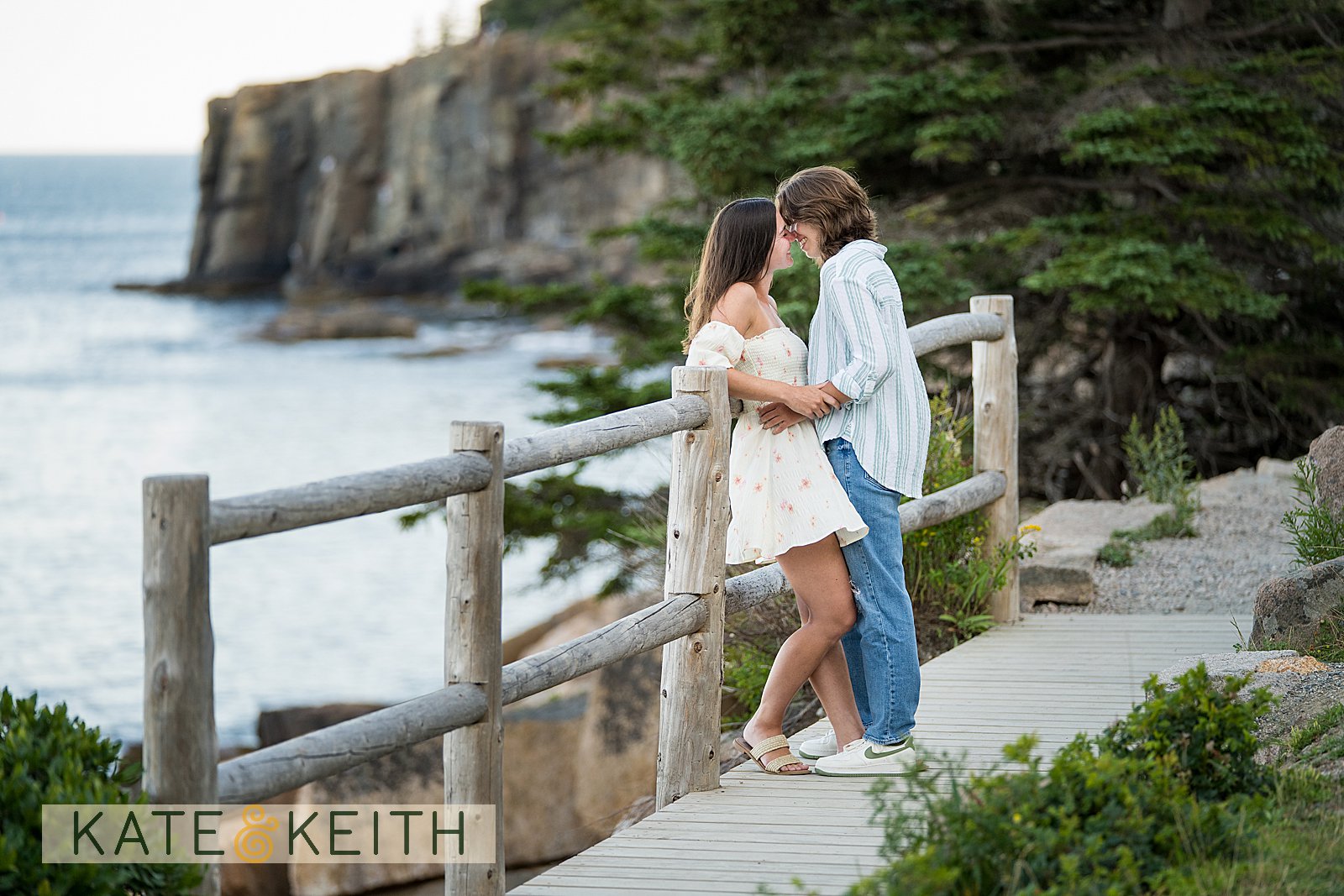 the cliffs of Acadia National Park in the background, a couple has a moment together leaning against a fence along the coastal path