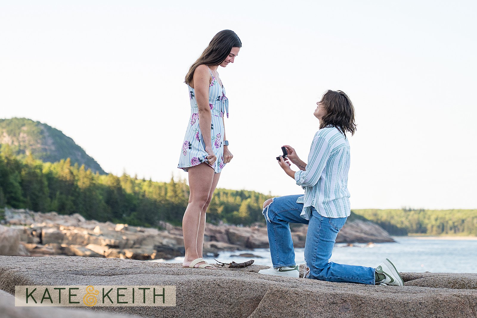 woman on one knee proposing to her girlfriend in Acadia National Park, with mountains and ocean in the background