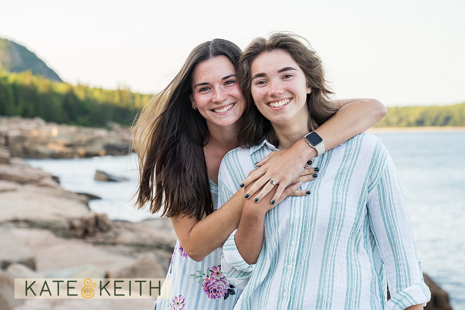 a couple standing with arms around each other, showing off an engagement ring on the coastline of Acadia National Park