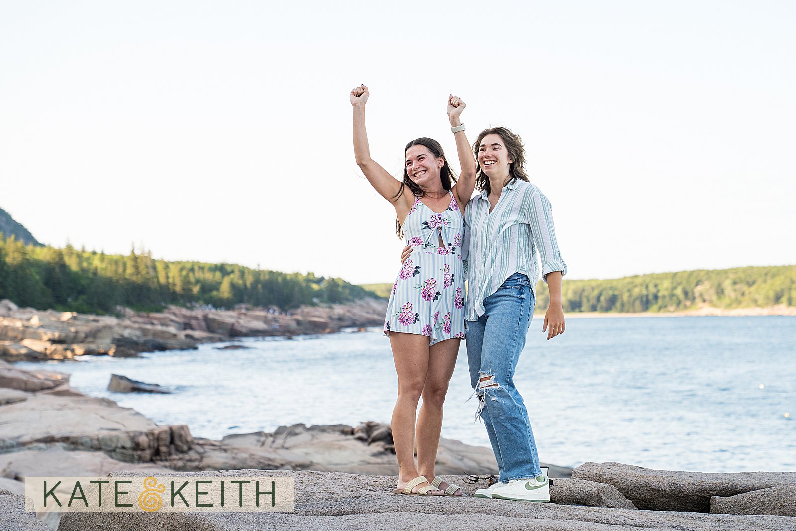 two women cheering after their proposal in Acadia National Park, with the Maine coast behind them