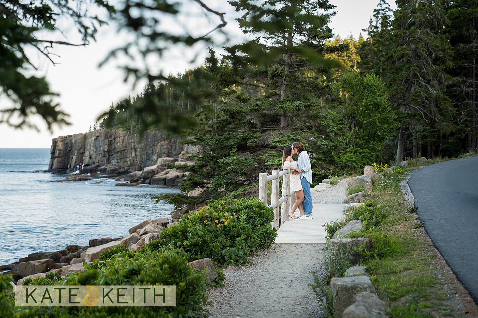 the cliffs of Acadia National Park in the background, a couple has a moment together leaning against a fence along the coastal path