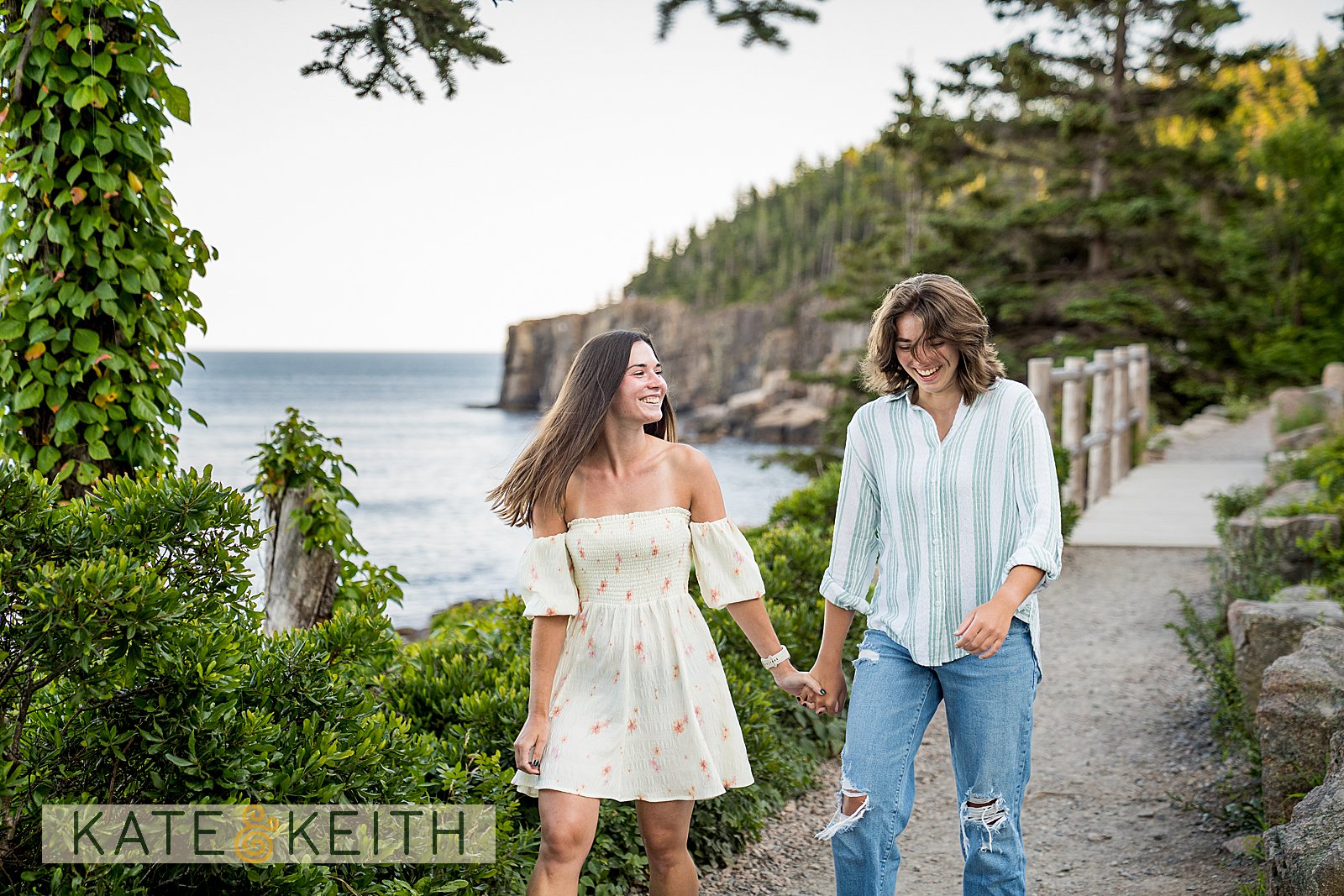 the cliffs of Acadia National Park in the background, a couple walks holding hands along a coastal path