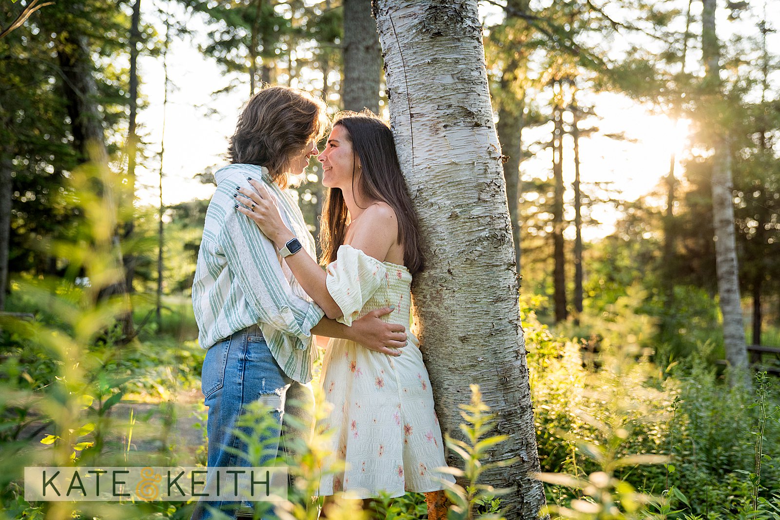 a couple leaning against a tree embracing among ferns with the setting sun behind them